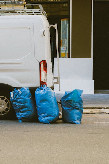 A collection of black garbage bags piled on the ground in front of a red metal door set into a textured, off-white exterior wall of a building. Some of the bags are tied and bulging, indicating they a