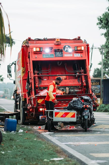 A waste collection worker, wearing an orange high-visibility vest and dark trousers, is positioned behind a large red rubbish truck with the rear loading mechanism open. The worker is actively engaged