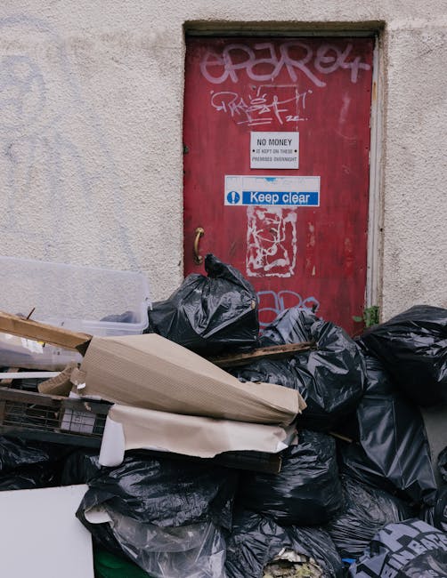 A waste collection worker, wearing an orange high-visibility vest and dark trousers, is positioned behind a large red rubbish truck with the rear loading mechanism open. The worker is actively engaged