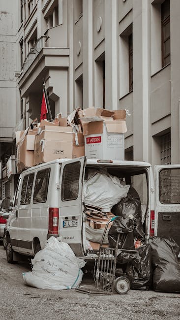 A sanitation worker wearing a high-visibility yellow and red uniform is performing rubbish collection on a public street, standing beside a large red waste collection truck with its rear hopper open, 